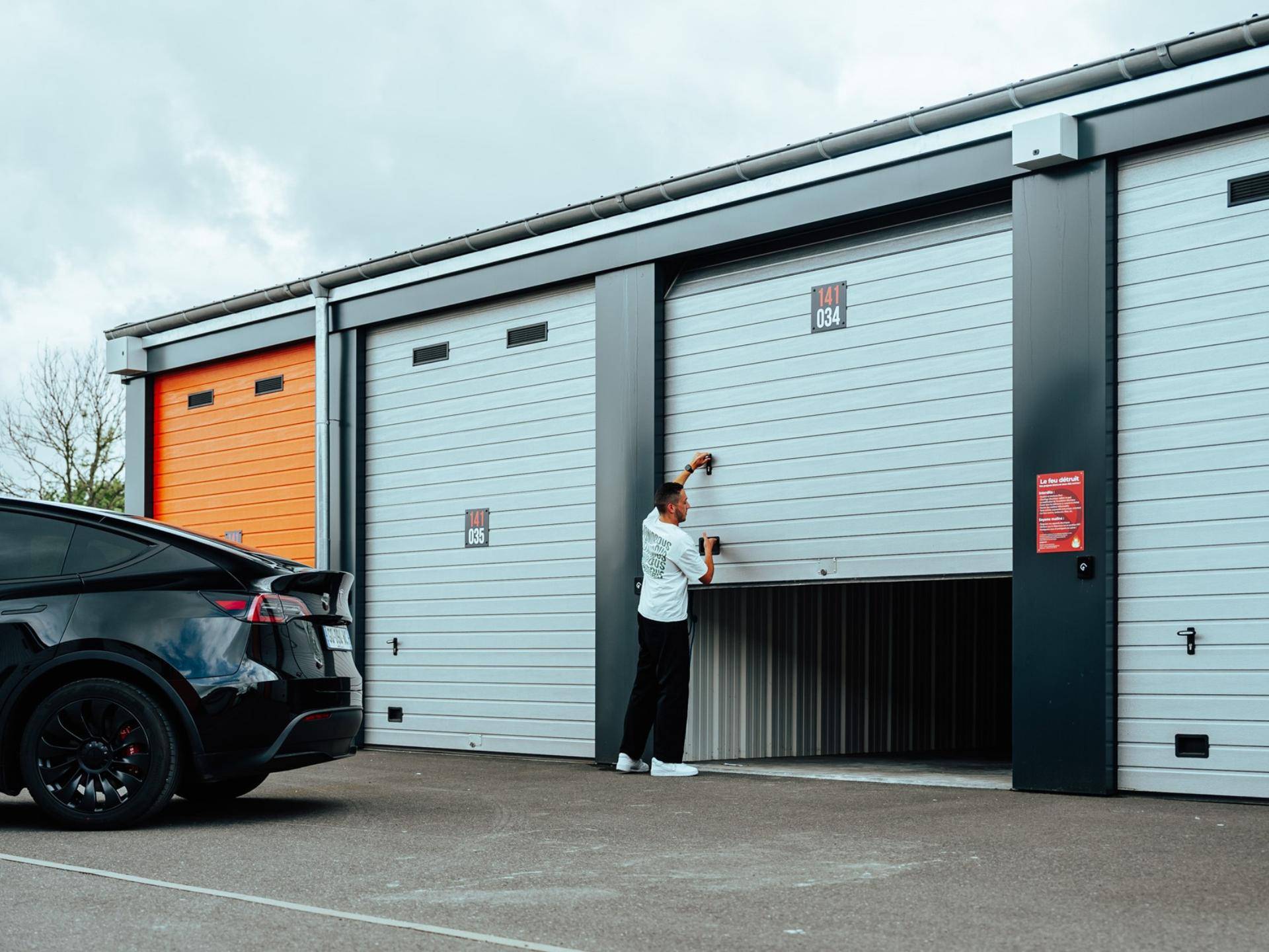 Eine Person öffnet das Rolltor einer Lagerhalle, während ein schwarzes Auto davor parkt.