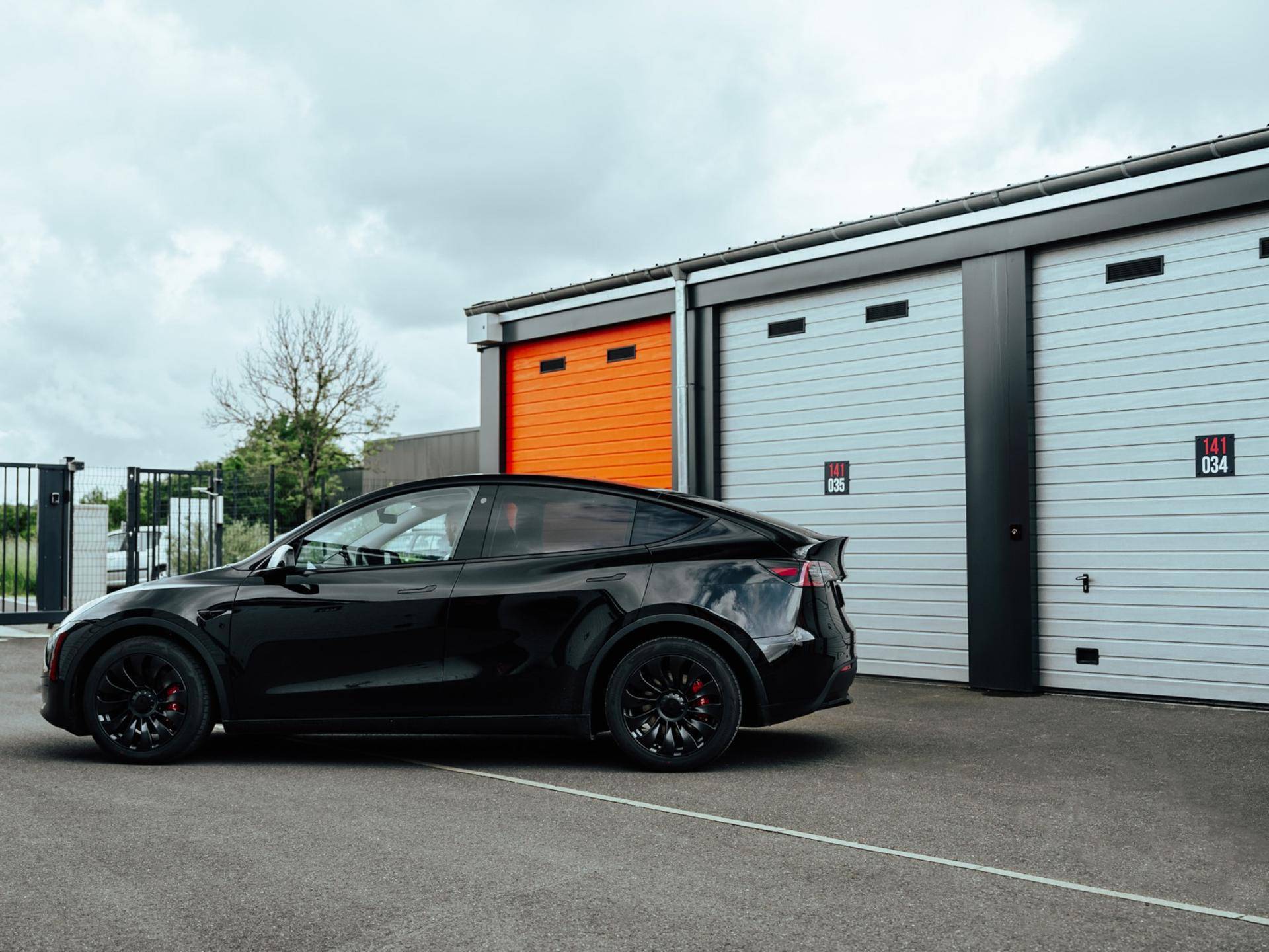 Schwarzes Auto parkt vor modernen Lagerhallen mit grauen und orangefarbenen Rolltoren.
