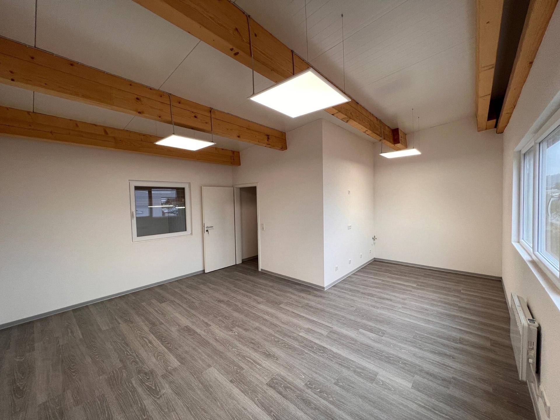 Heller Büroraum mit Holzbalken, großen Fenstern und modernem Vinylboden in einem Neubau.
