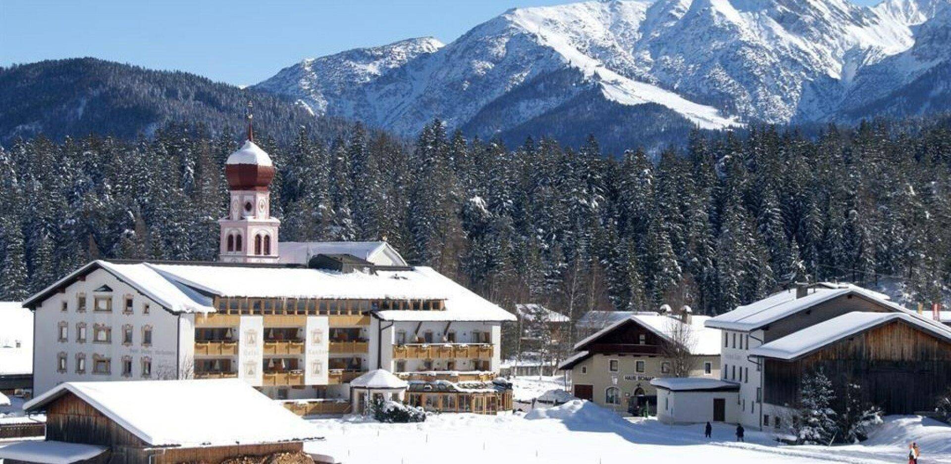 Winterliche Ansicht eines Hotels und umliegender Gebäude vor einer beeindruckenden Bergkulisse mit verschneiten Gipfeln.