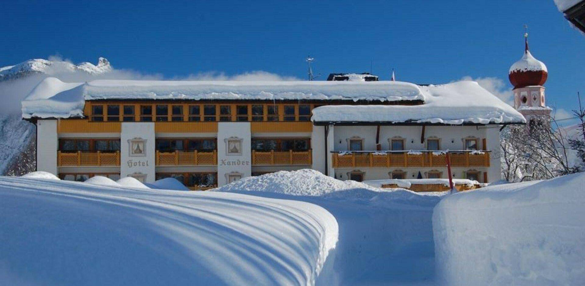 Das Hotelgebäude ist im Winter tief verschneit, mit sichtbaren Balkonen und einem klaren blauen Himmel über den Bergen.