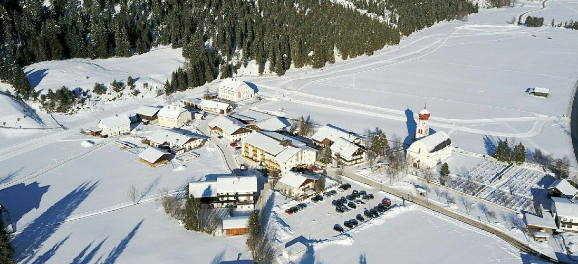 Luftaufnahme eines verschneiten Dorfes mit Häusern, Wald und einer Kirche in einer winterlichen Berglandschaft.