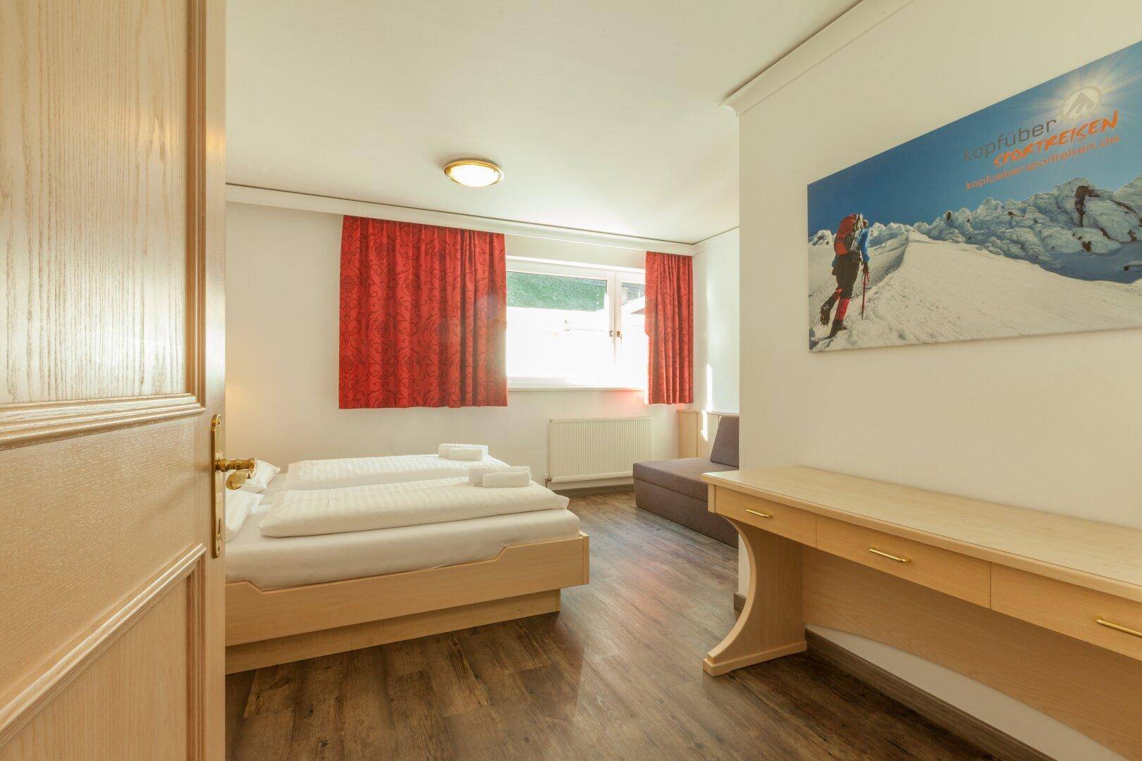 Helles Schlafzimmer mit Doppelbett, Schreibtisch und Laminatboden, beleuchtet durch ein Fenster.
