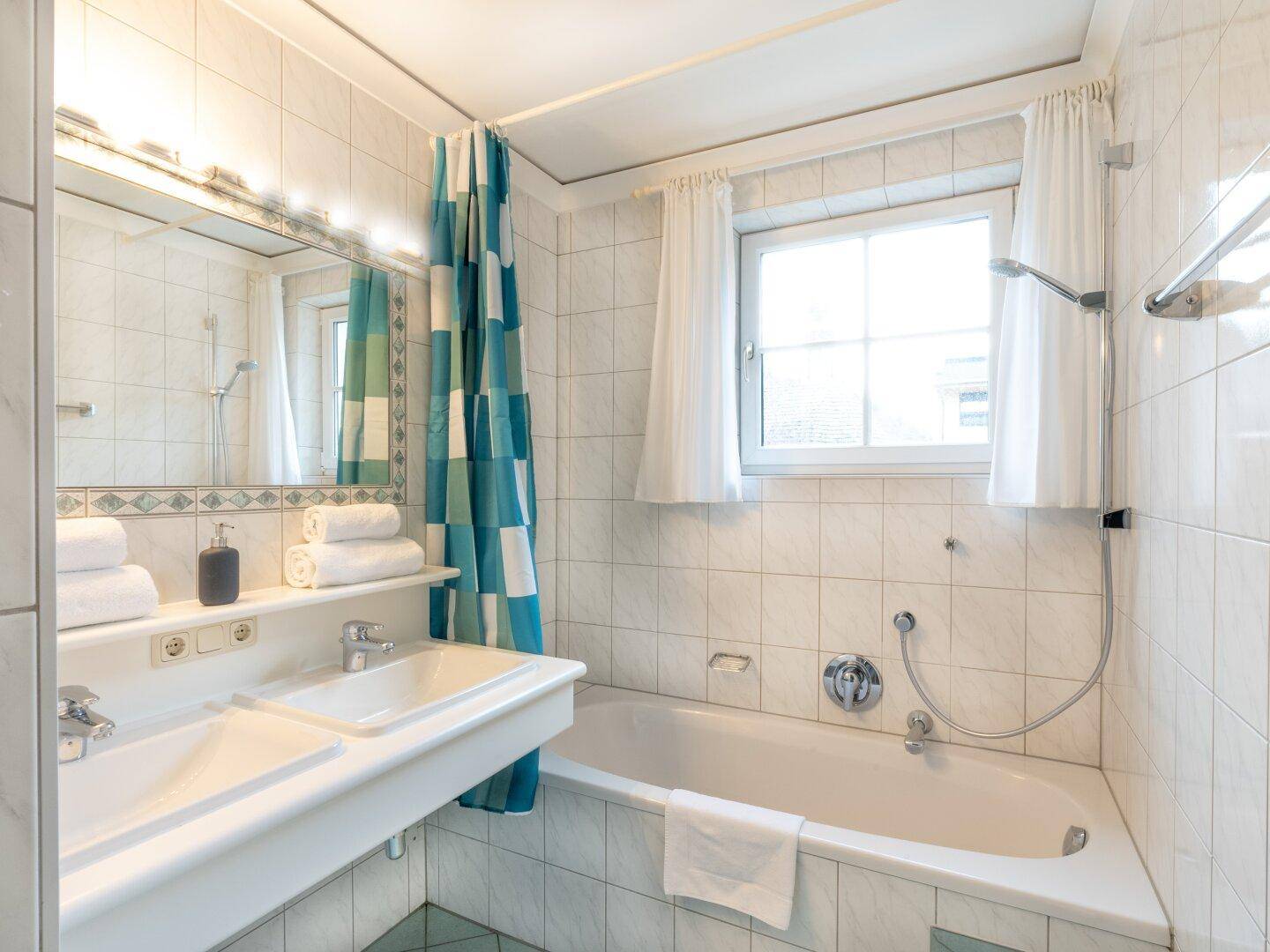 Helles Badezimmer mit Badewanne, Duschvorrichtung und Fenster, gefliest in Weiß und Blau.