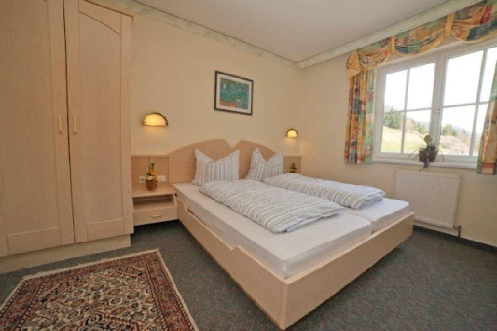 Gemütliches Schlafzimmer mit Doppelbett, Kleiderschrank und Teppichboden, hell durch Fenster.