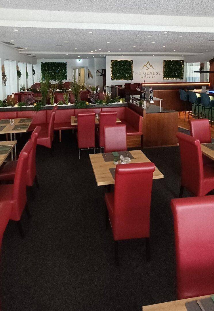 Weitläufiger Restaurantraum mit roten Stühlen und Bänken, sowie einer Bar im Hintergrund.