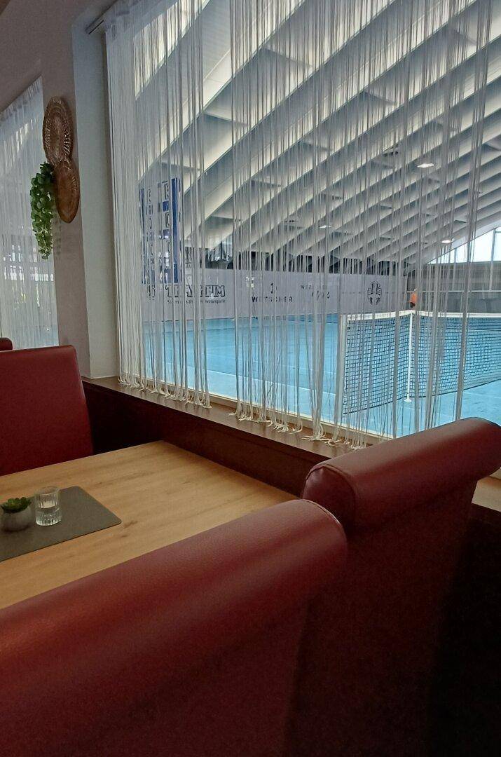 Restaurantbereich mit Blick durch große Fenster auf eine Tennishalle.