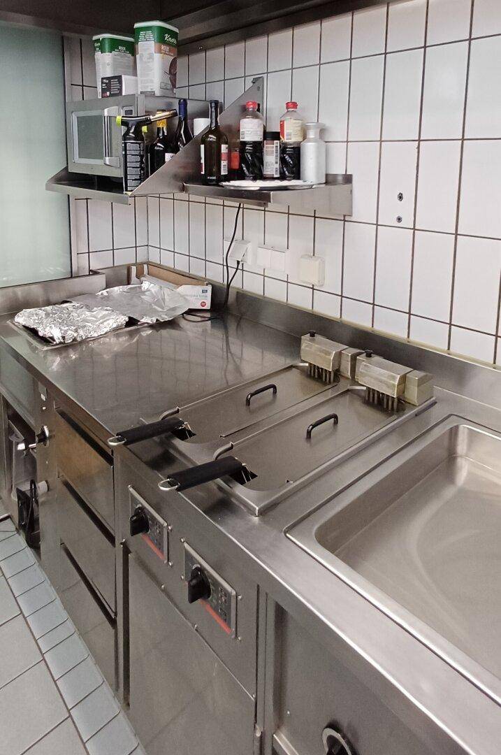 Arbeitsbereich in einer Großküche mit Fritteusen, Edelstahlflächen und Wandregal.