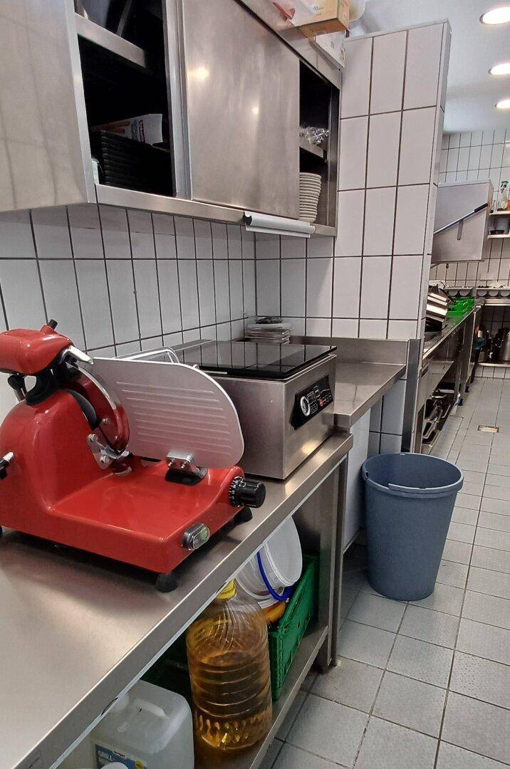 Küchenarbeitsplatz mit Aufschnittmaschine, Induktionskochfeld und Edelstahlmöbeln.