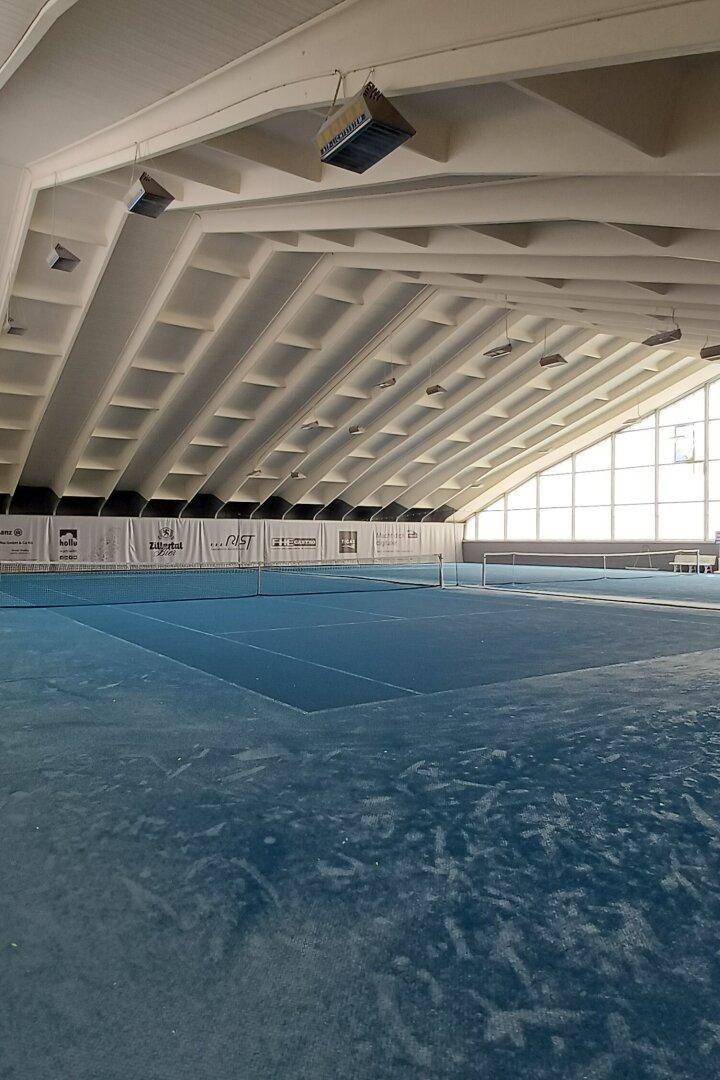 Große Tennishalle mit blauem Boden, hohen Decken und natürlichem Lichteinfall.