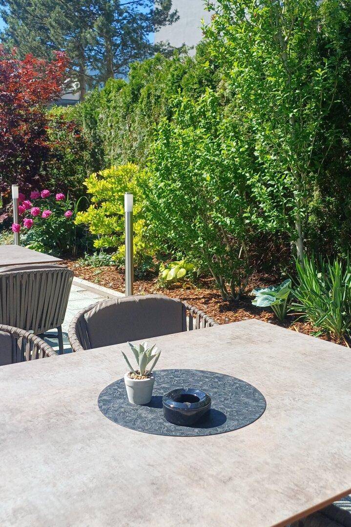 Detailaufnahme eines Tisches auf der Terrasse mit üppiger grüner Bepflanzung im Hintergrund.