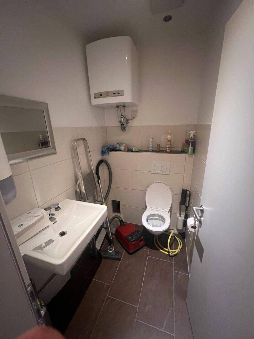 Einfache Toilette mit Waschbecken, Wand-WC und Warmwasserbereiter, mit Fliesen an Wänden und Boden.