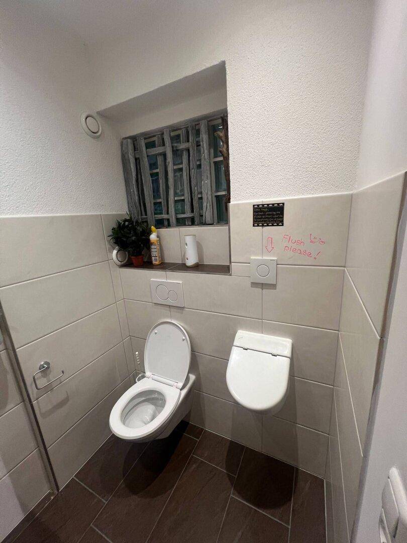 Gästetoilette mit zwei Wand-WCs und einem kleinen Fenster, gefliesten Wänden und dunklem Fliesenboden.