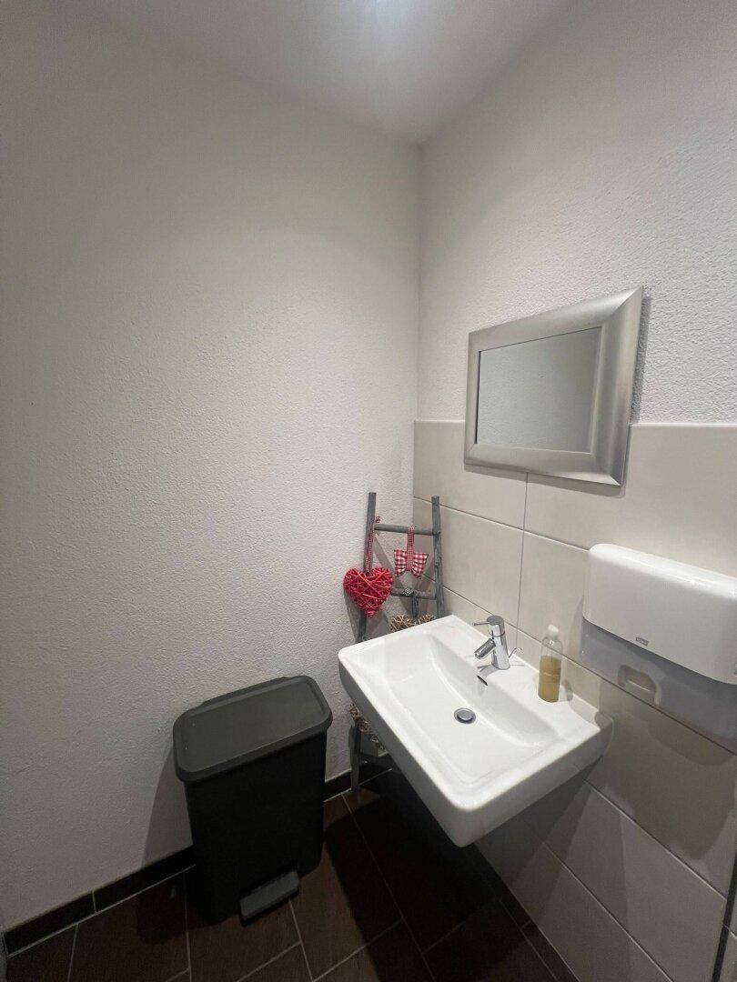 Waschbereich in einer Toilette mit Waschbecken, Spiegel und Seifenspender, auf dunklem Fliesenboden.