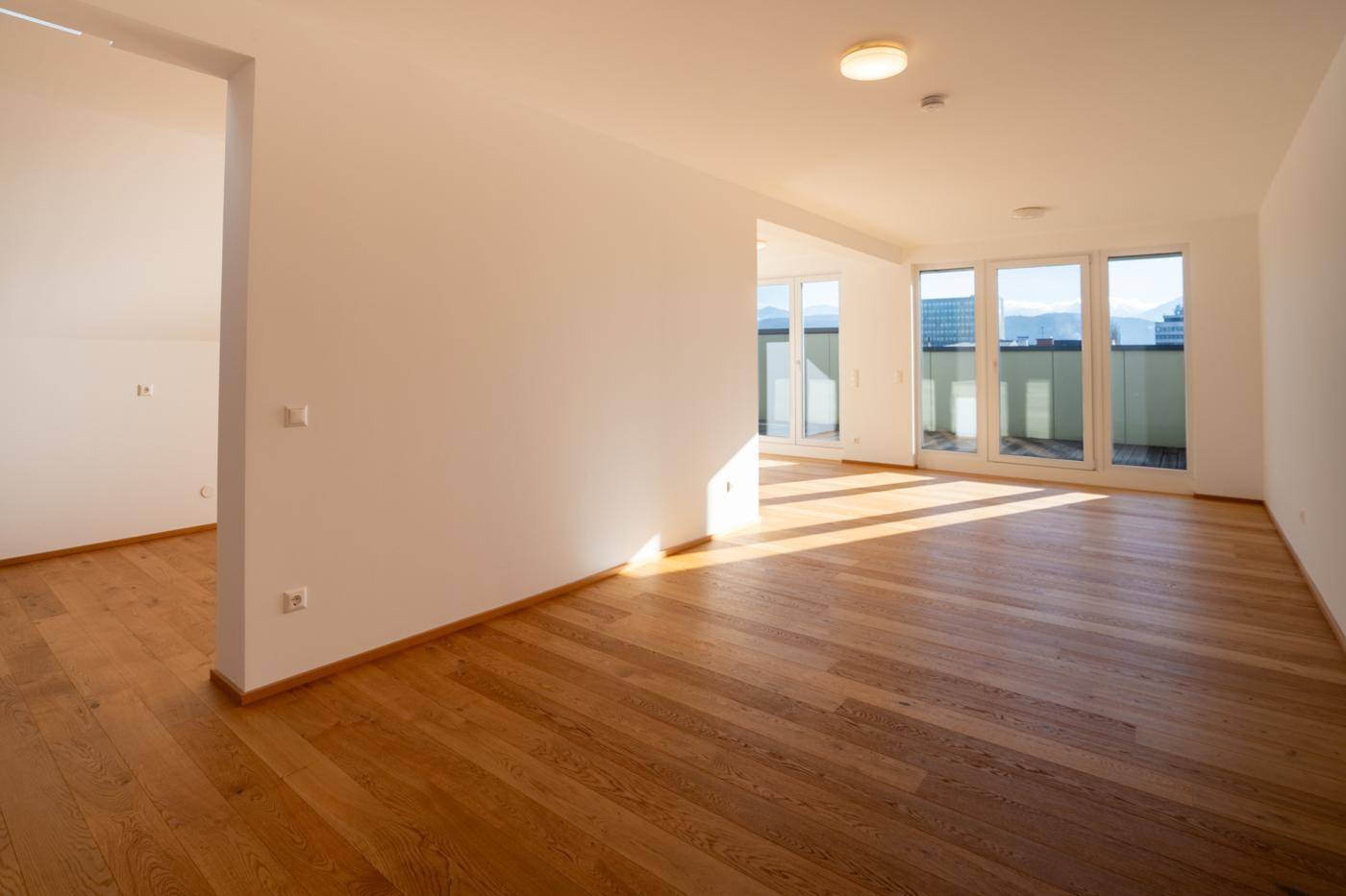 Leeres, lichtdurchflutetes Wohnzimmer mit Holzboden und großen Fenstern, die Zugang zur Terrasse bieten.