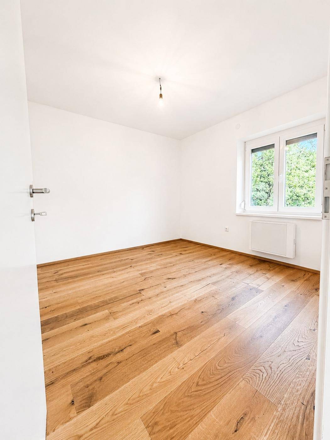 Leeres Schlafzimmer mit hellem Holzboden und einem Fenster mit Blick ins Grüne.