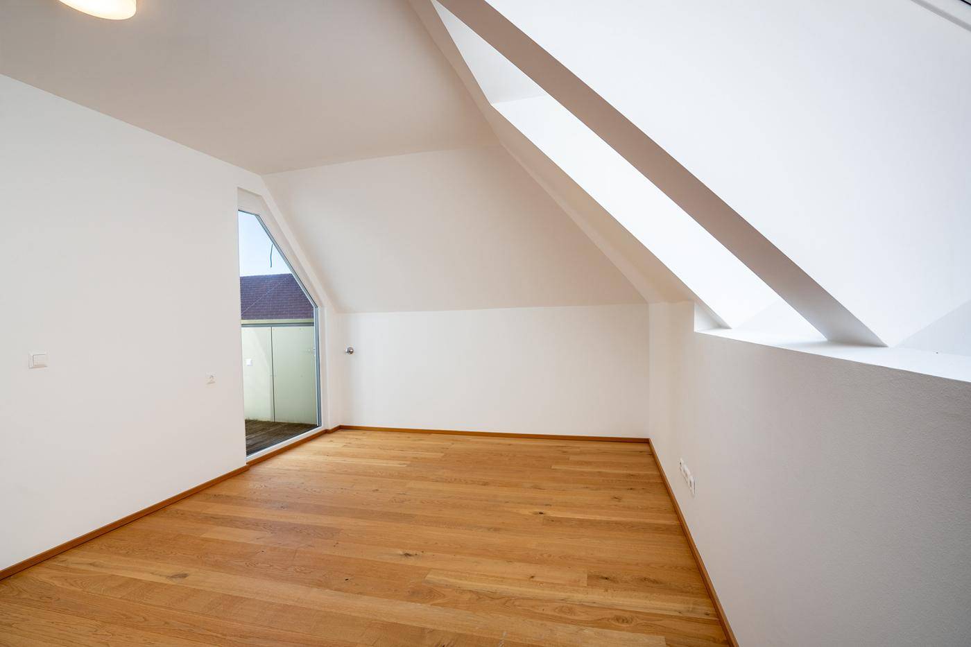 Heller, leerer Raum mit Holzboden, Dachschrägen und Zugang zu einem Außenbereich.