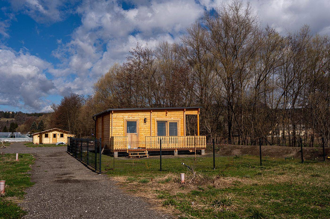 Kleines Holzhaus mit Zaun und Schotterweg, umgeben von Bäumen und Grünflächen.