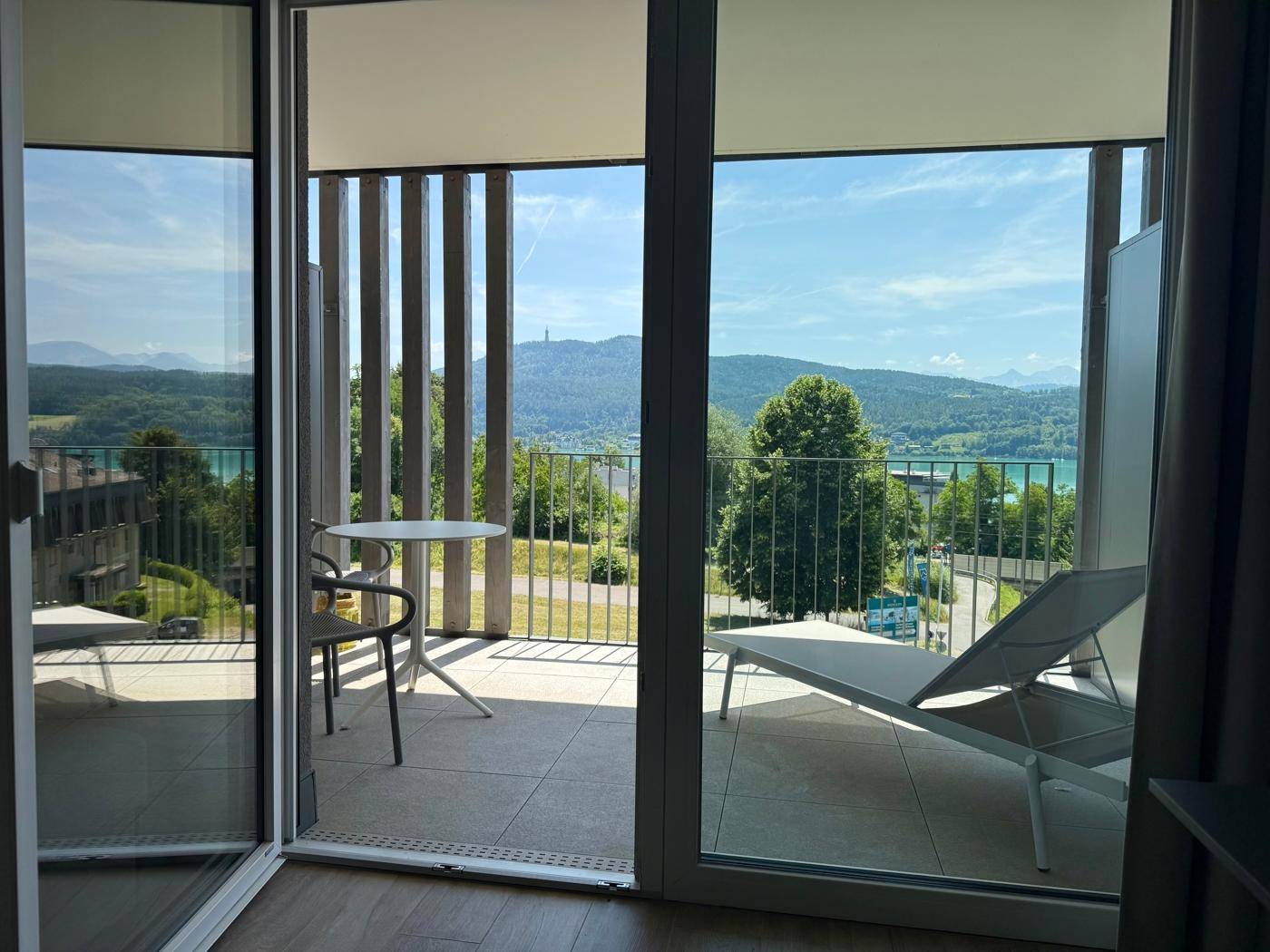 Blick vom Innenraum auf den Balkon mit Sitzgelegenheiten und weitreichender See- und Bergsicht.
