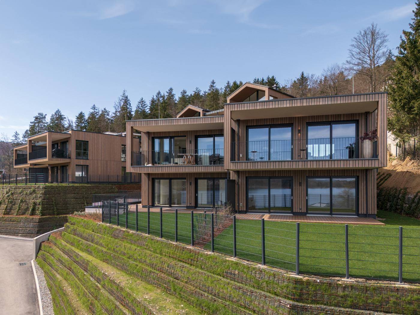 Moderne Wohngebäude mit Holzfassaden und großen Fensterfronten in Hanglage mit Seeblick.