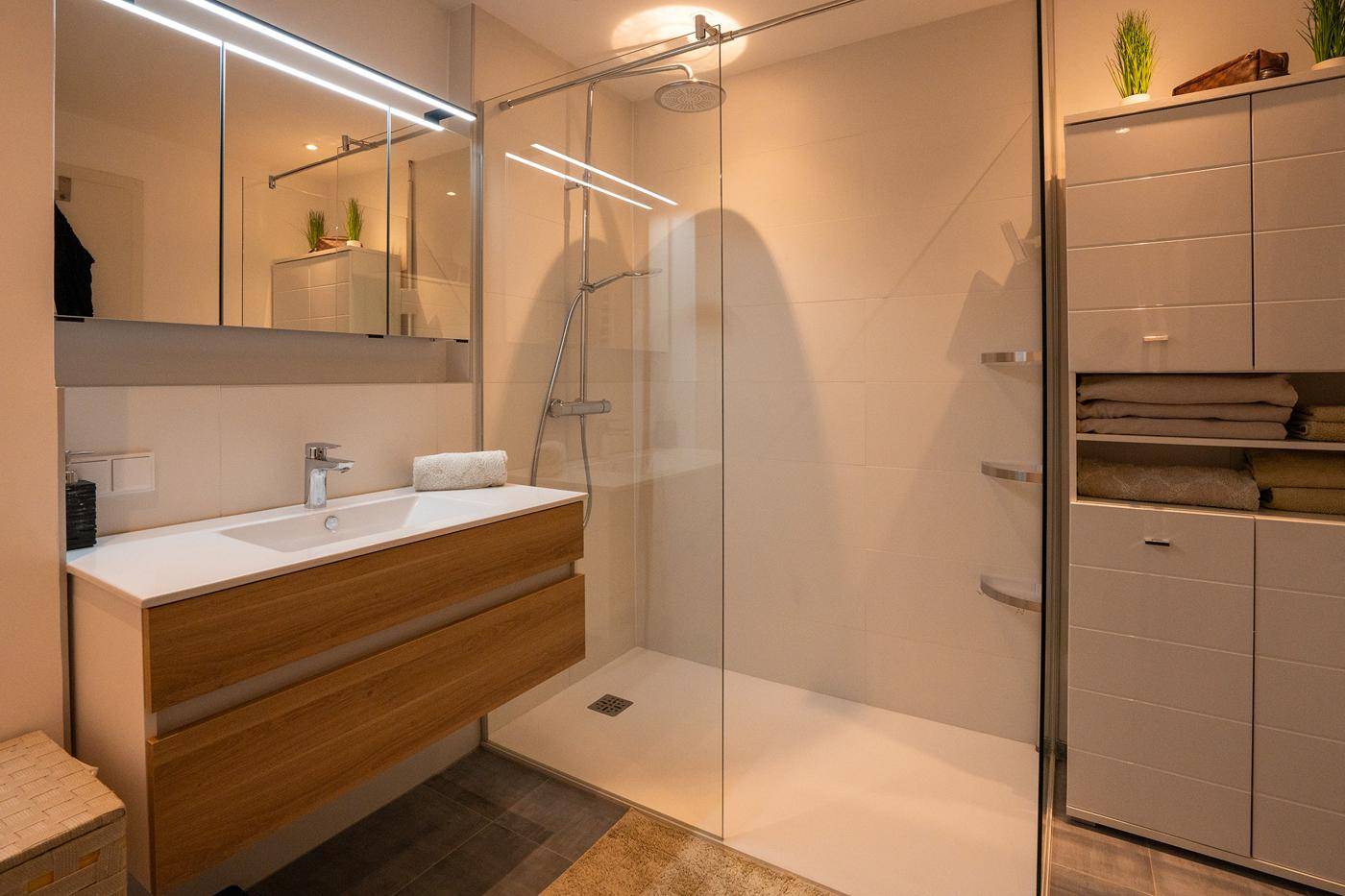 Modernes Badezimmer mit begehbarer Dusche, Holzwaschtisch und beleuchtetem Spiegelschrank.