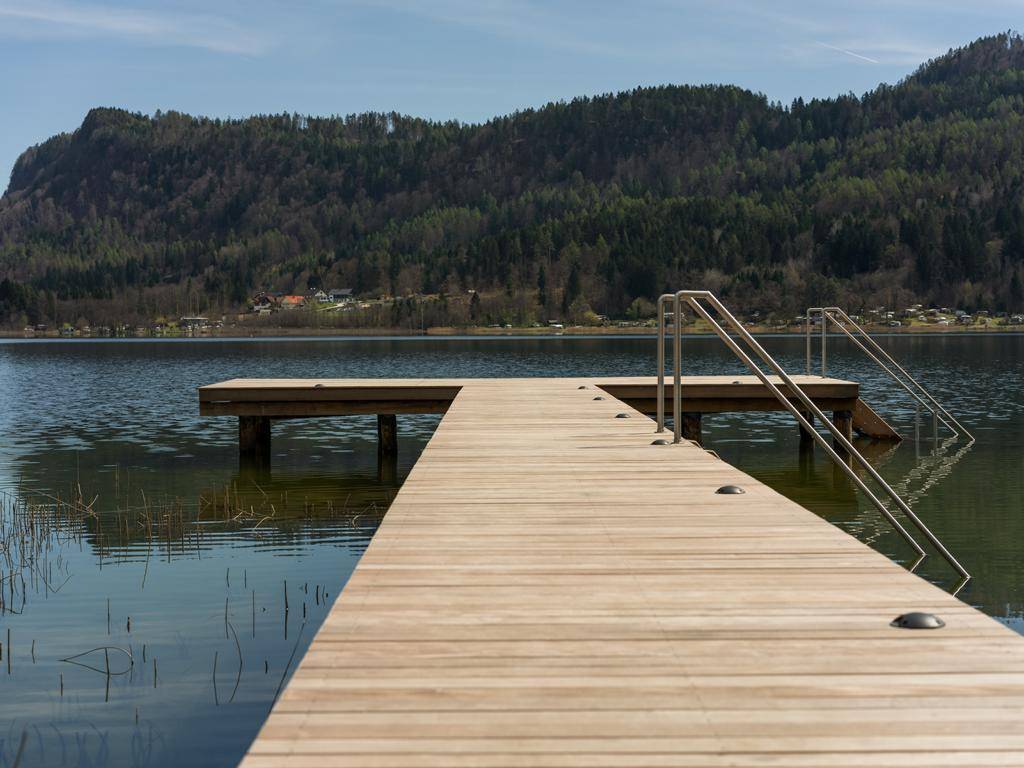 Langer Holzsteg ragt in den ruhigen See, umgeben von bewaldeten Hügeln unter blauem Himmel.