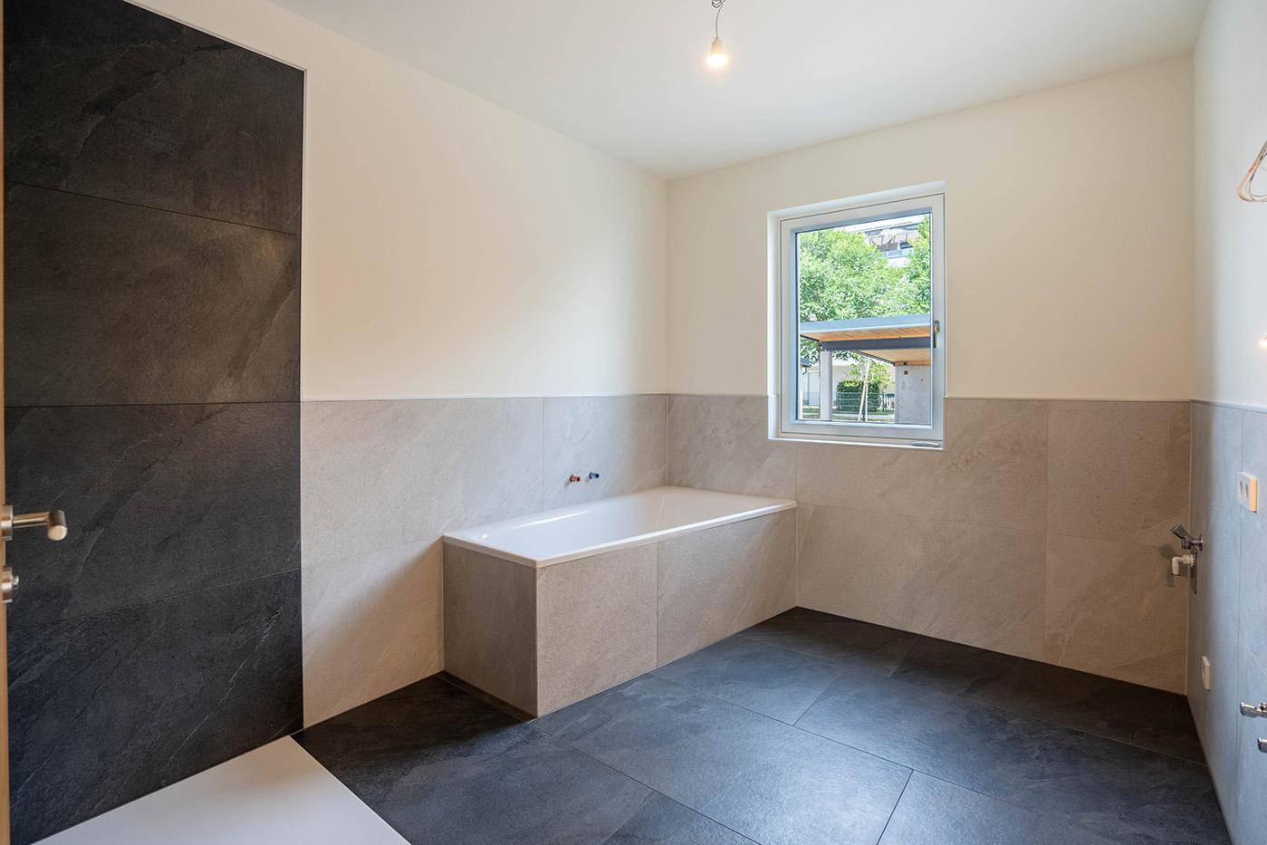 Modernes Badezimmer mit Badewanne, großformatigen Fliesen und Fenster für Tageslicht.