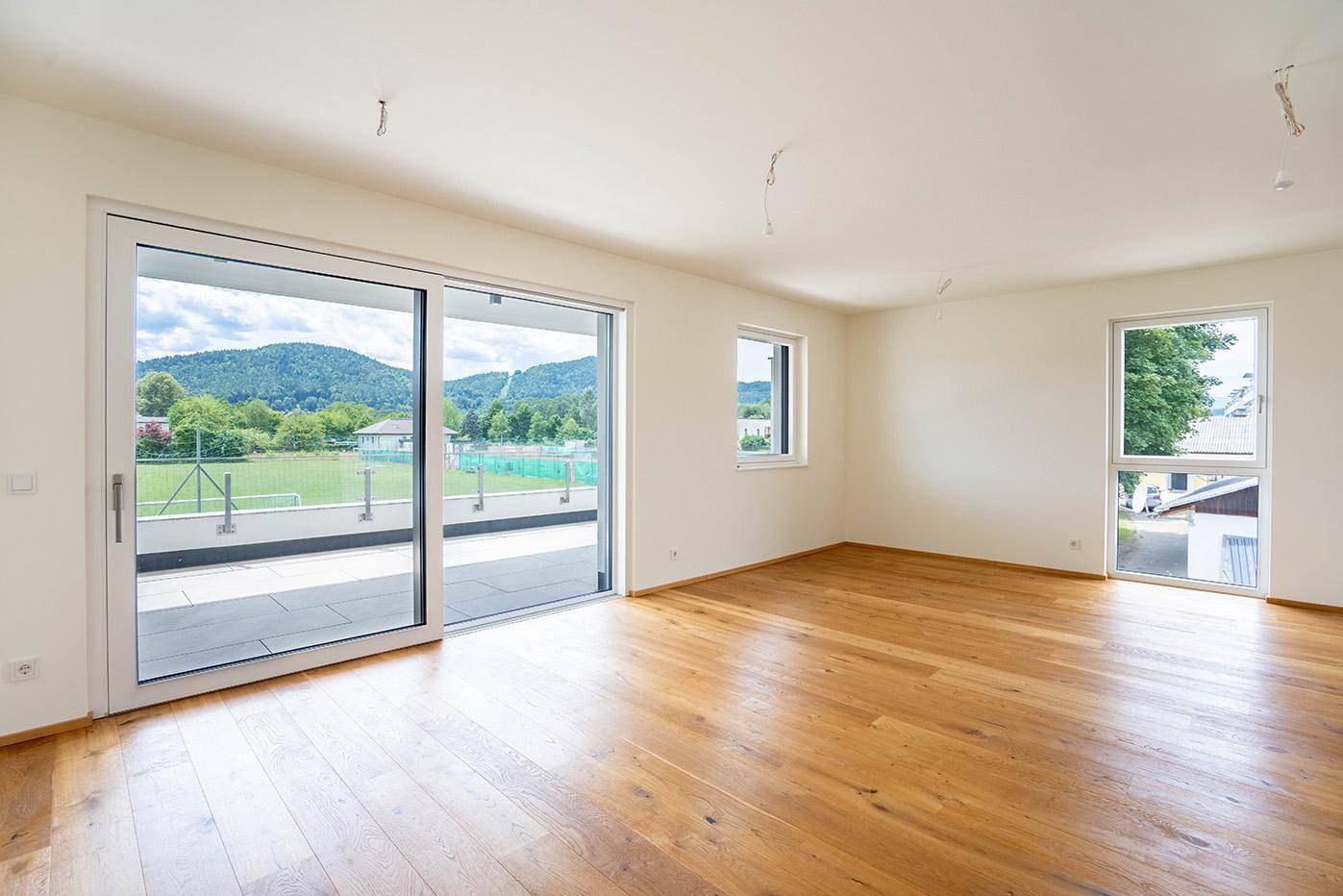 Helles, leeres Zimmer mit Holzboden und großen Schiebetüren, die auf einen Balkon mit Bergblick führen.