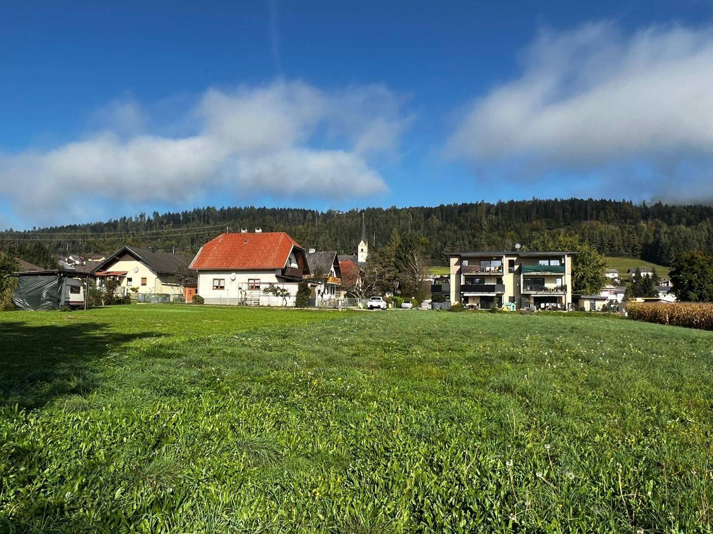 Weitläufige grüne Wiese mit Blick auf ein Dorf und bewaldete Hügel im Hintergrund.