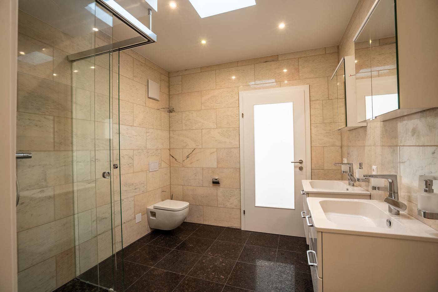 Modernes Badezimmer mit doppelten Waschbecken, begehbarer Dusche, Wand-WC und Oberlicht für Tageslicht.