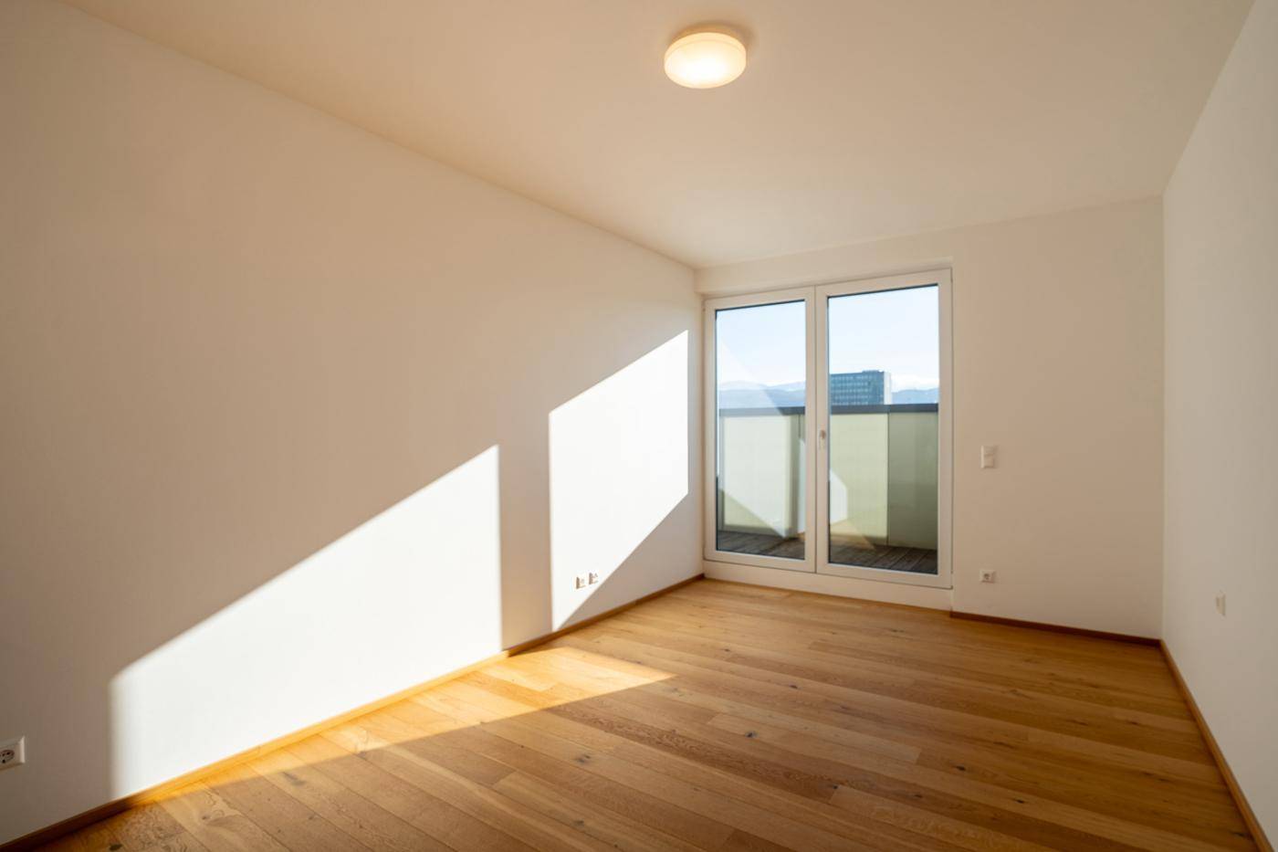 Sonnenverwöhntes Zimmer mit Holzboden und Zugang zu einem kleinen Balkon, ideal als Schlaf- oder Gästezimmer.