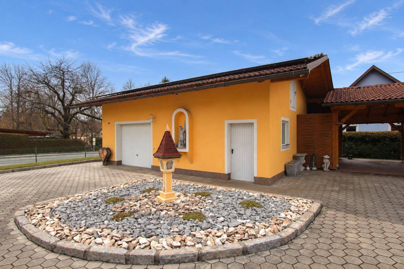 Separate Garage mit gelber Fassade, gepflastertem Vorplatz und dekorativem Kiesbeet.