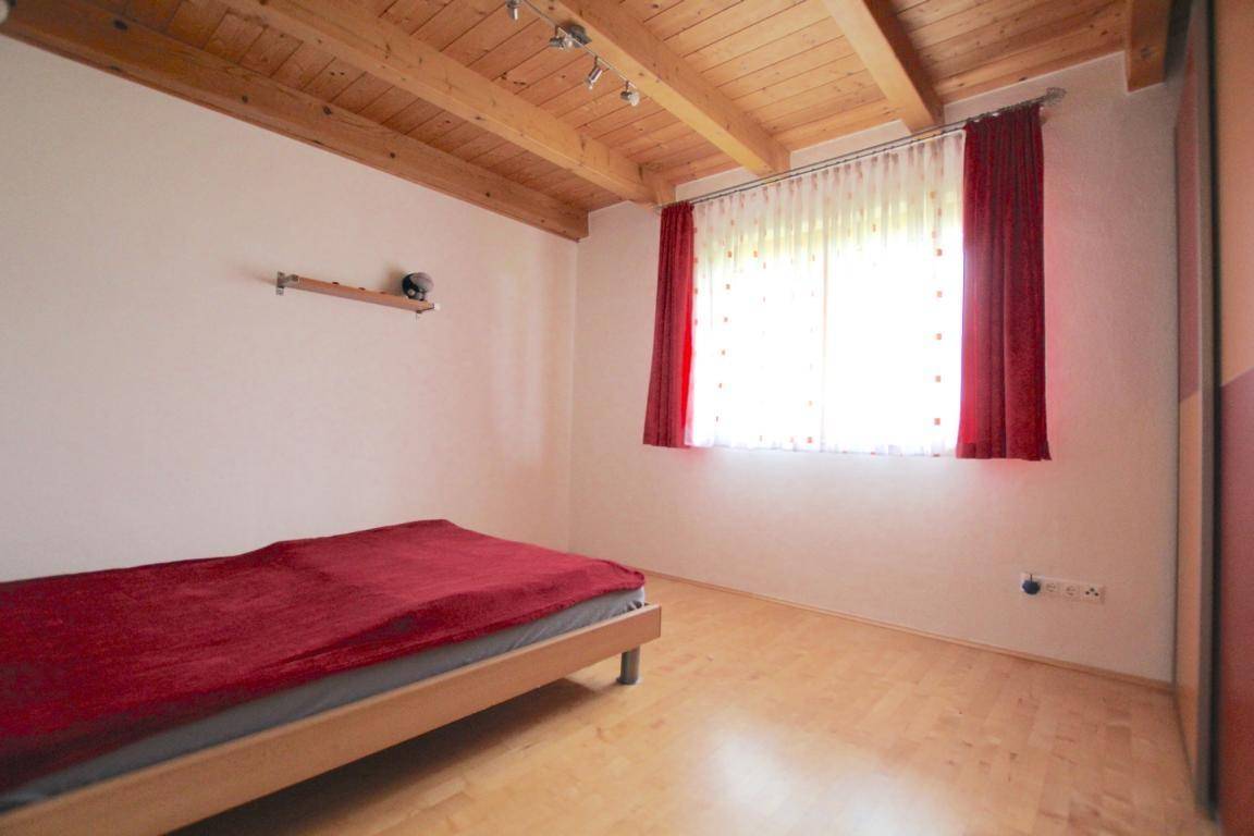 Schlafzimmer mit Holzbalkendecke, hellem Holzboden und rotem Bettüberwurf.