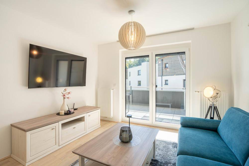 Gemütliches Wohnzimmer mit Sofa, Fernseher und großen Fenstern zum Balkon.