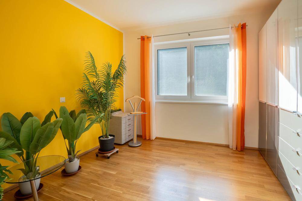 Helles Schlafzimmer mit gelber Akzentwand, Holzboden und großem Fenster.