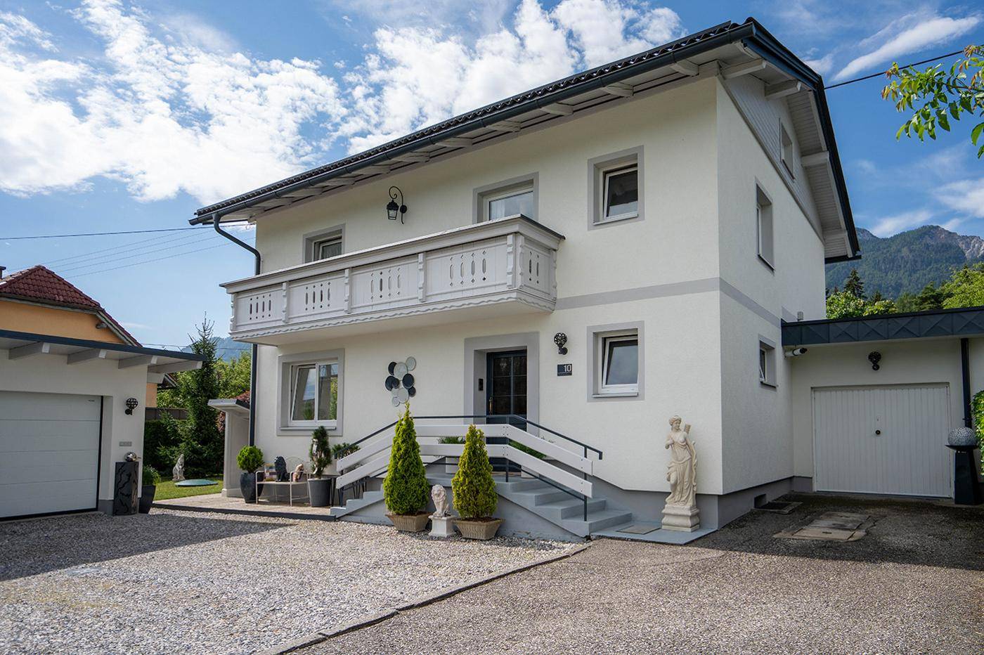 Vorderansicht des modernen Einfamilienhauses mit Balkon, gepflegtem Vorgarten und Blick auf die Berge.