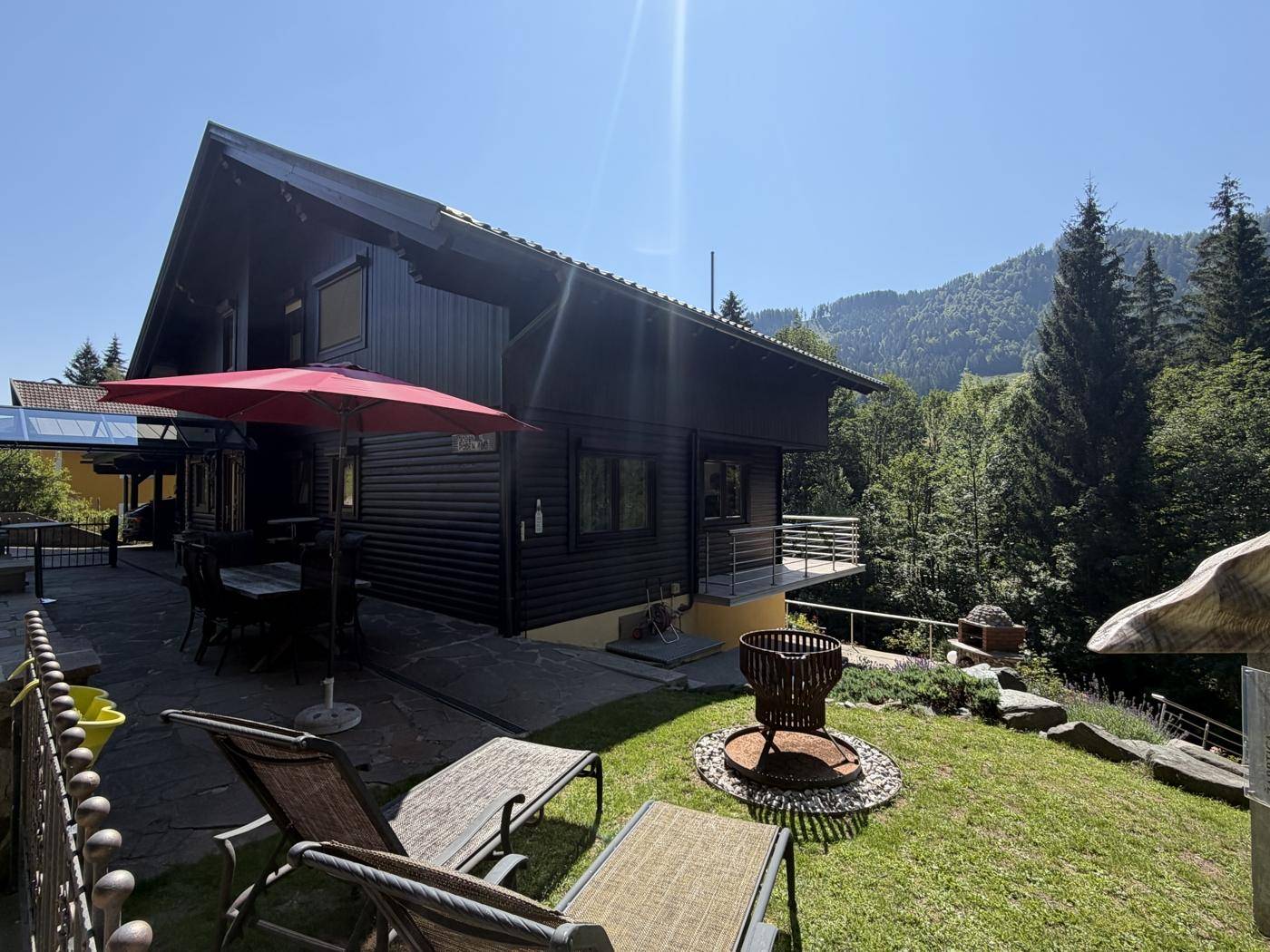 Sonniger Gartenbereich mit Liegestühlen, Feuerstelle und Blick auf die umliegenden Berge.