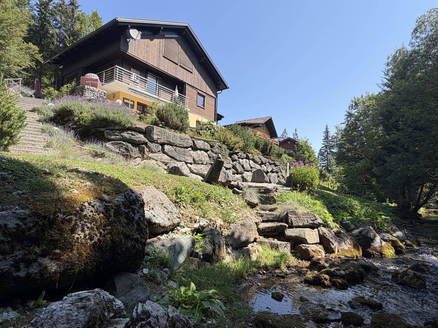 Charmantes Haus am Bachlauf, umgeben von üppiger Natur und felsigen Gartenbereichen.