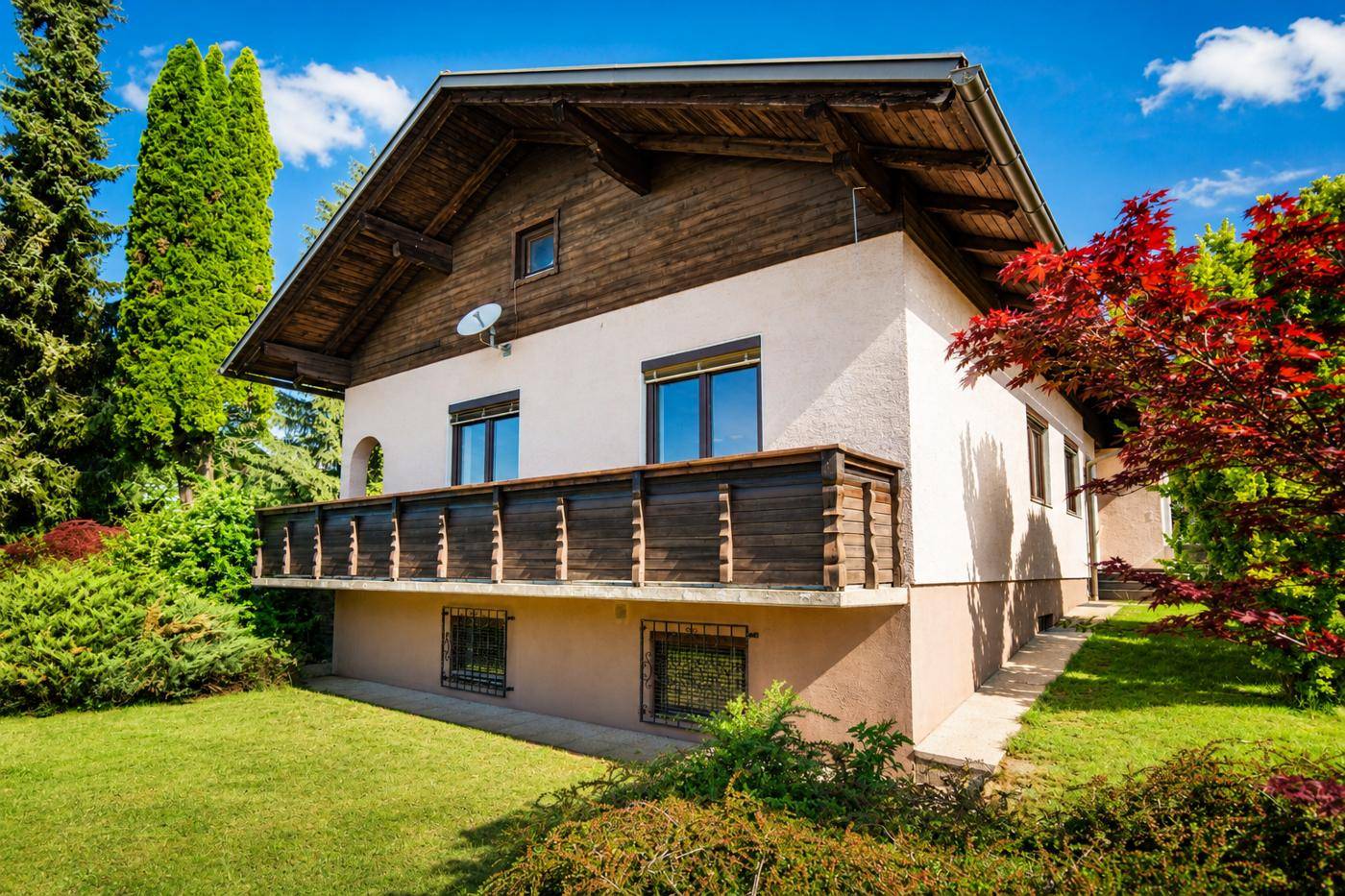 Seitenansicht des Hauses mit Holzbalkon, gepflegtem Rasen und hohen Bäumen.