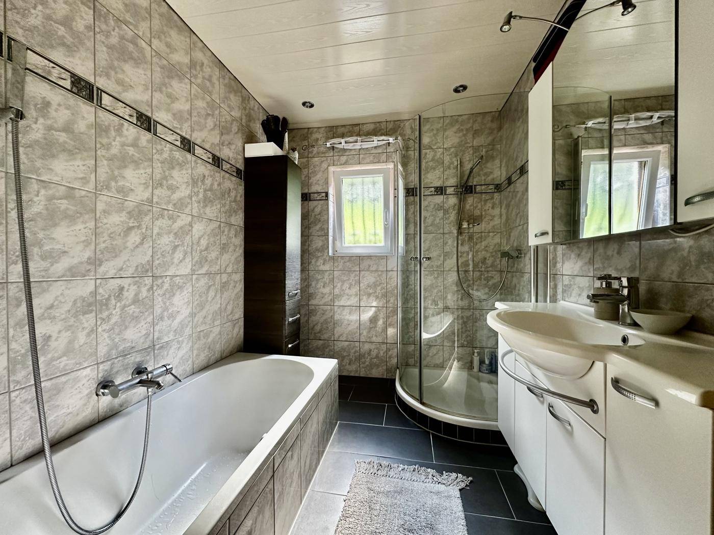 Gefliestes Badezimmer mit Badewanne, Dusche und Fenster, in neutralen Grautönen gehalten.