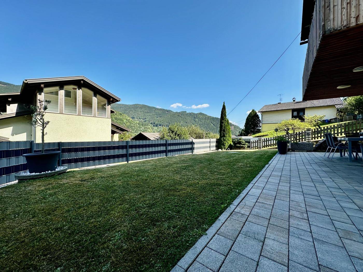 Großer, gepflegter Garten mit Rasenfläche und gepflasterter Terrasse, umgeben von einem Zaun und Bergblick.