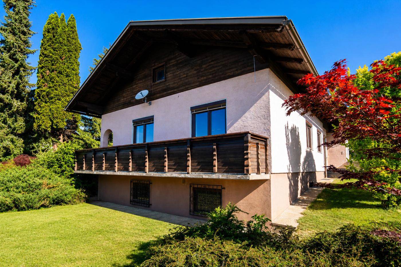 Ansicht des Hauses von der Seite mit Holzbalkon, großem Garten und hohen Bäumen.