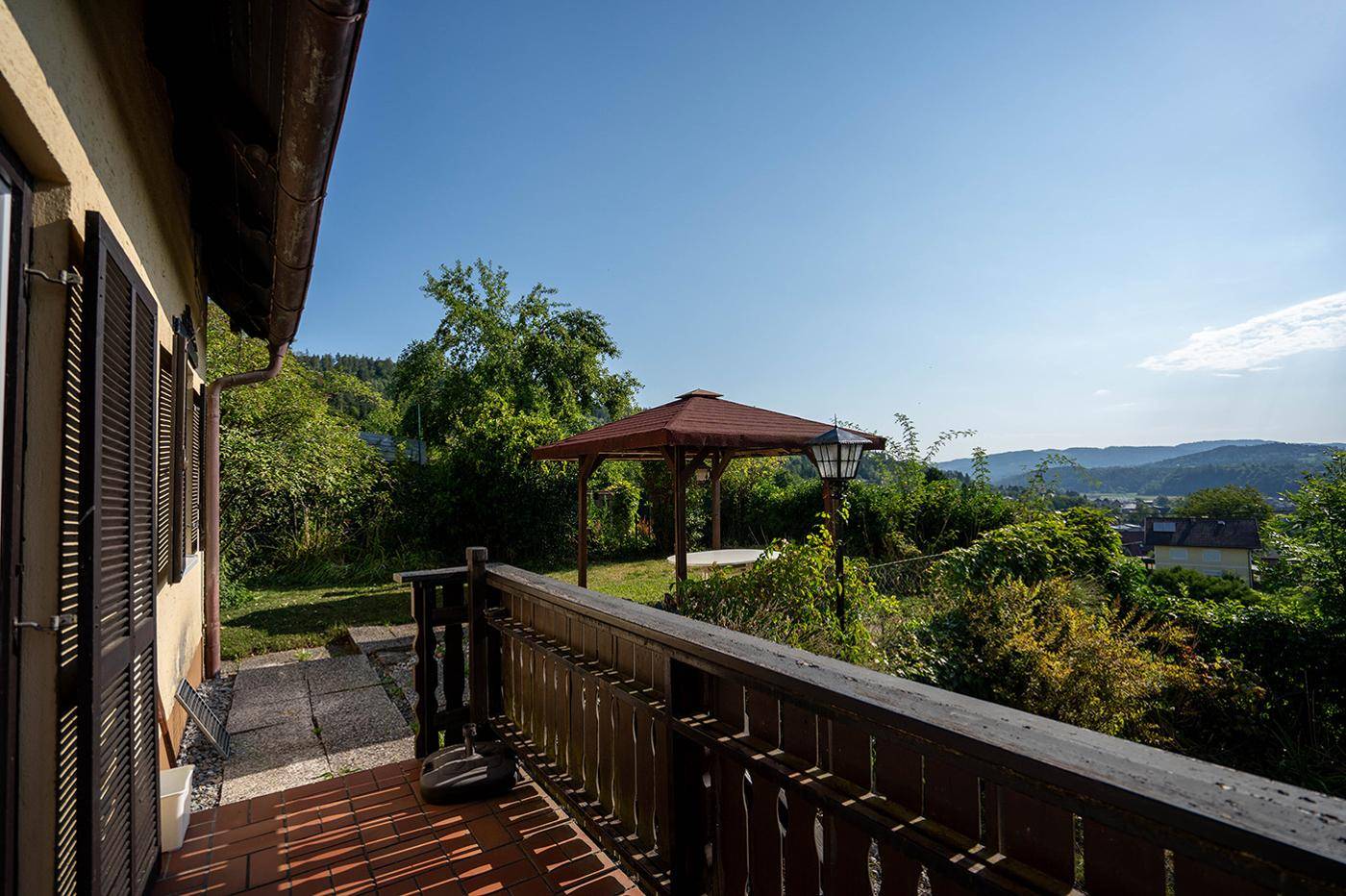 Sonniger Balkon mit Holzbalkon und weitem Blick über die grüne Landschaft und einen Pavillon.