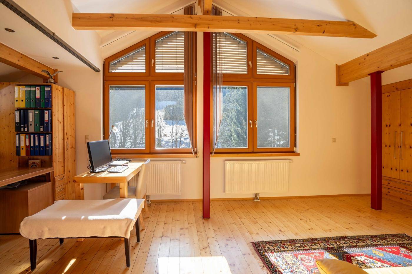 Helles Arbeitszimmer im Dachgeschoss mit Holzbalken, großen Fenstern und Waldblick.