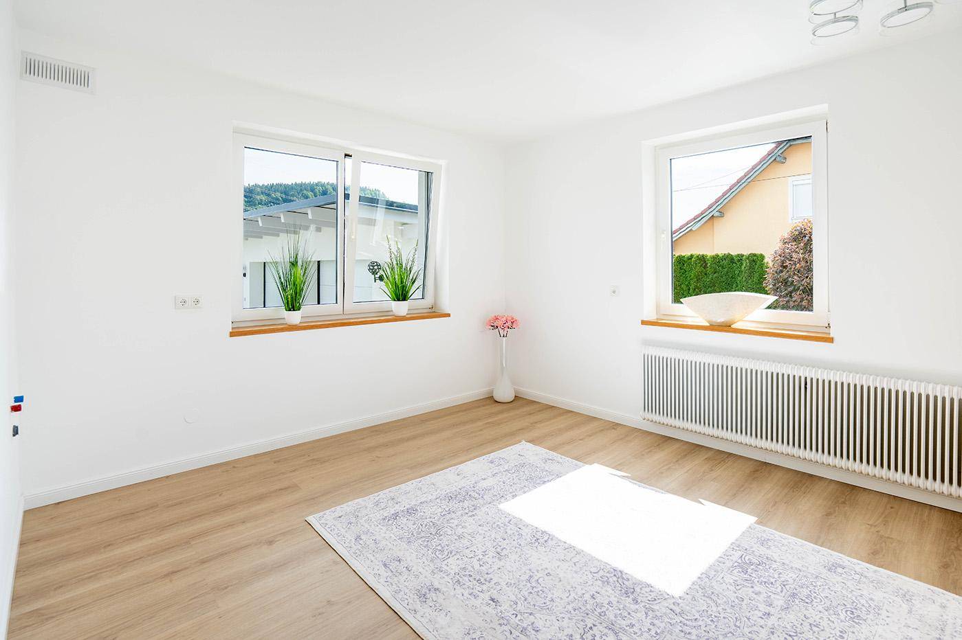 Heller Raum mit Laminatboden, zwei Fenstern und einem Heizkörper unter dem Fenster.