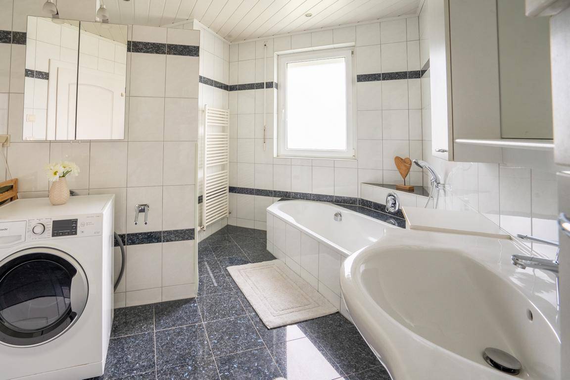 Gefliestes Badezimmer mit Badewanne, Waschbecken, Waschmaschine und Fenster.