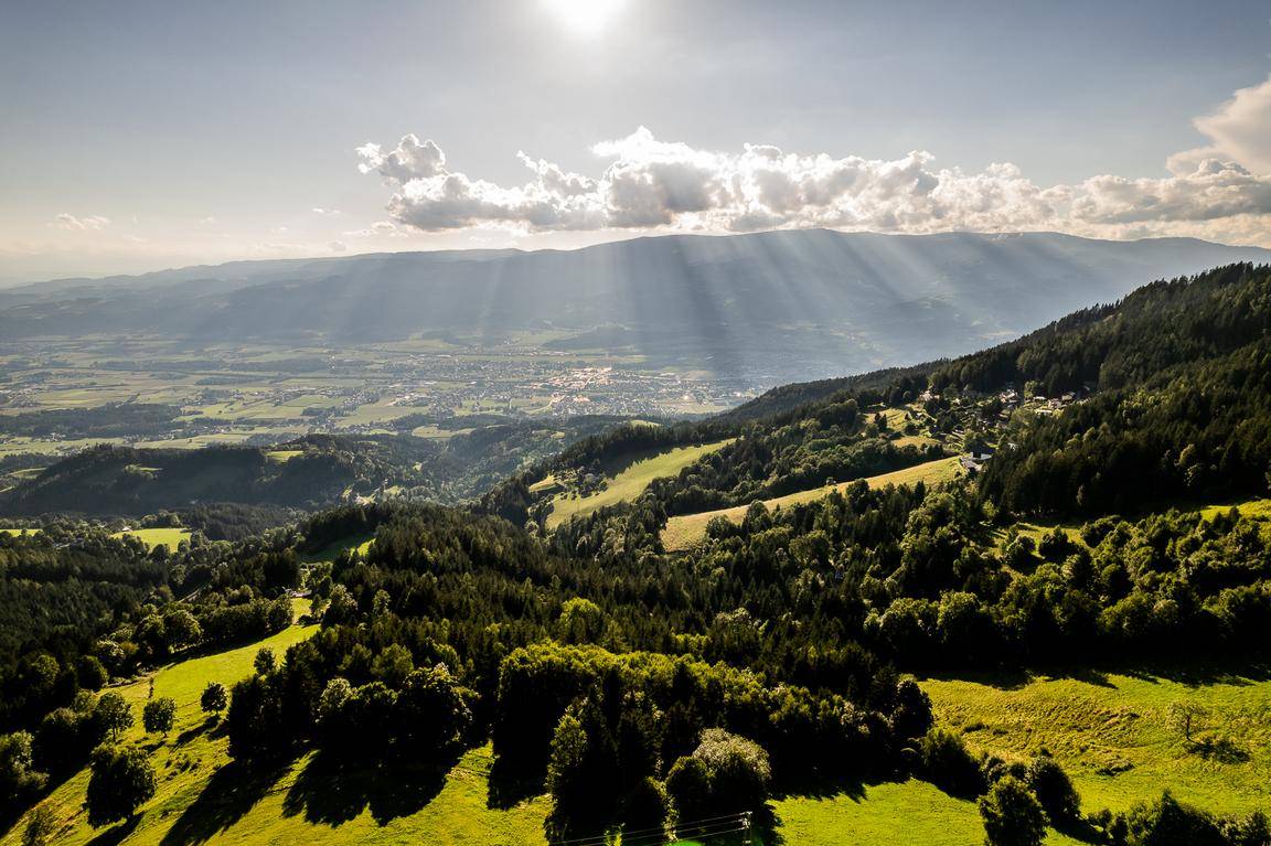 Panoramablick über ein weites Tal mit Wäldern, Bergen und einer Siedlung in der Ferne.