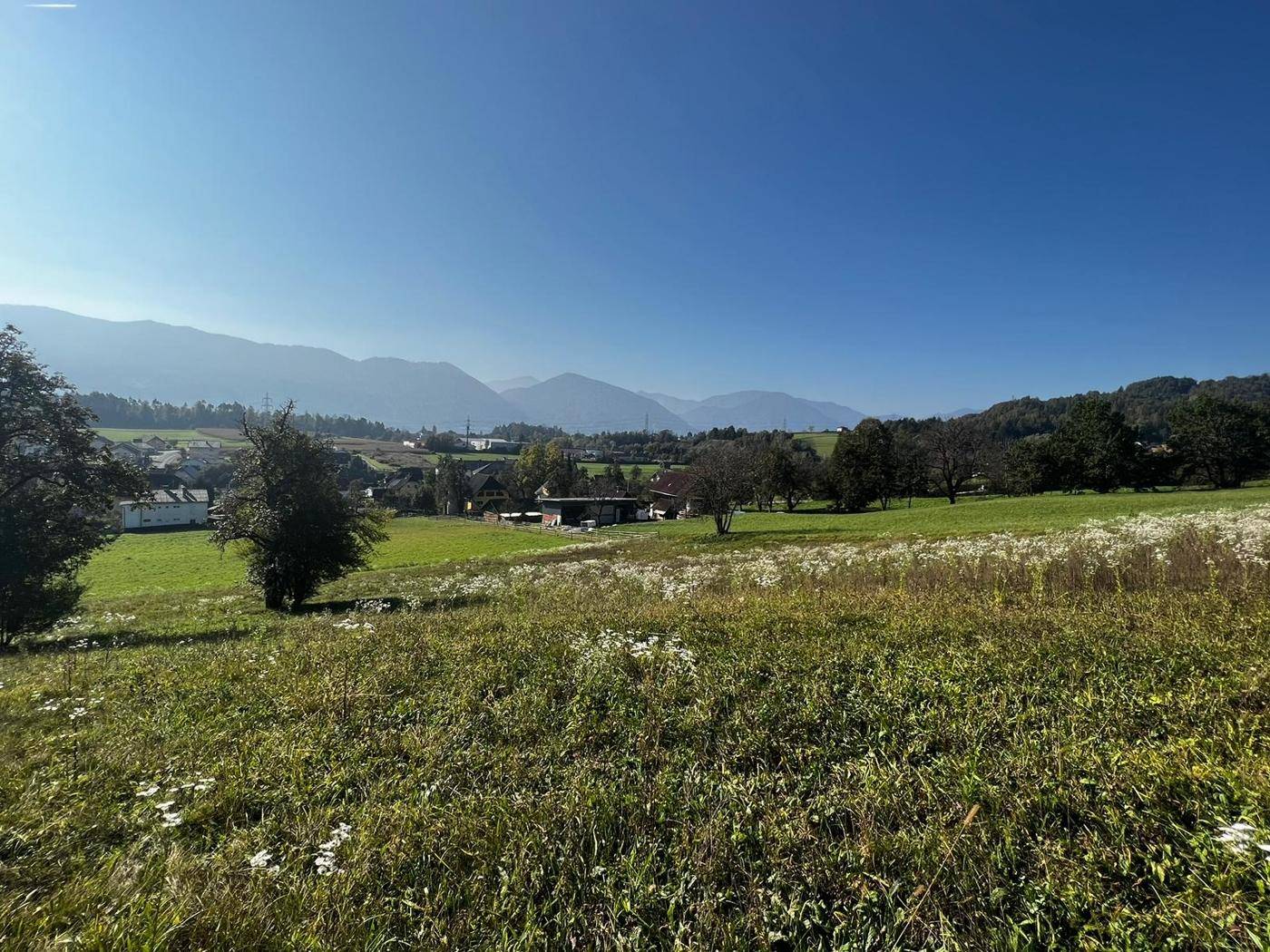 Weitläufige grüne Landschaft mit einem kleinen Dorf und einer beeindruckenden Bergkette am Horizont.
