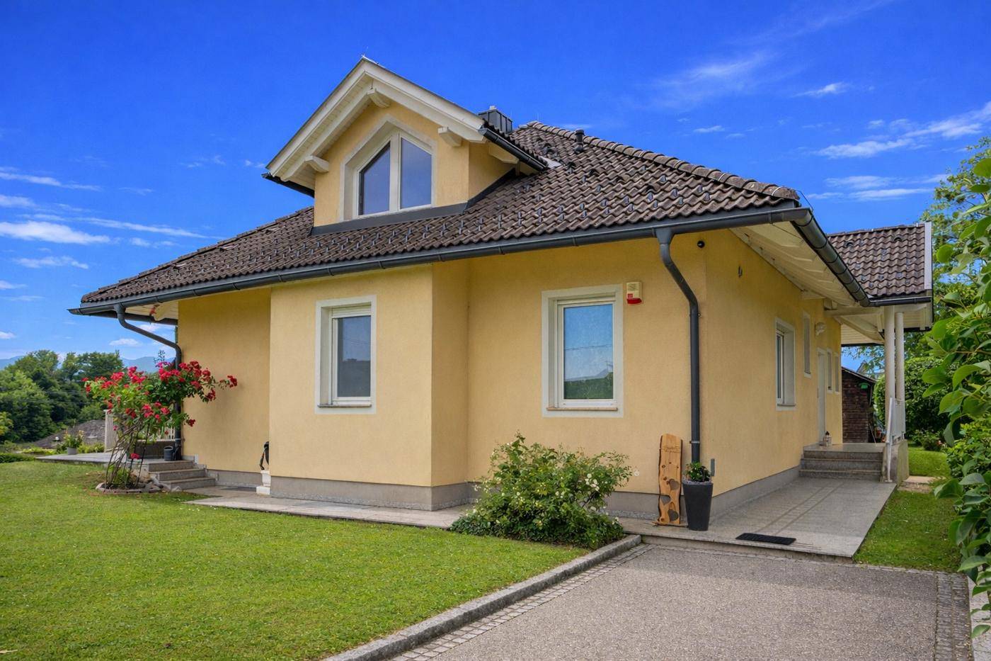 Seitenansicht des gelben Hauses mit gepflasterter Auffahrt und blühenden Pflanzen.