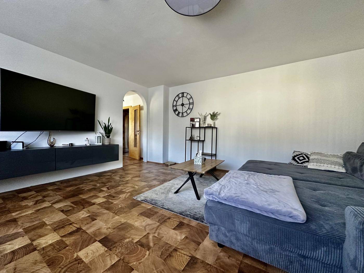 Helles Wohnzimmer mit Holzboden, großem Sofa und Wandfernseher, modern eingerichtet.